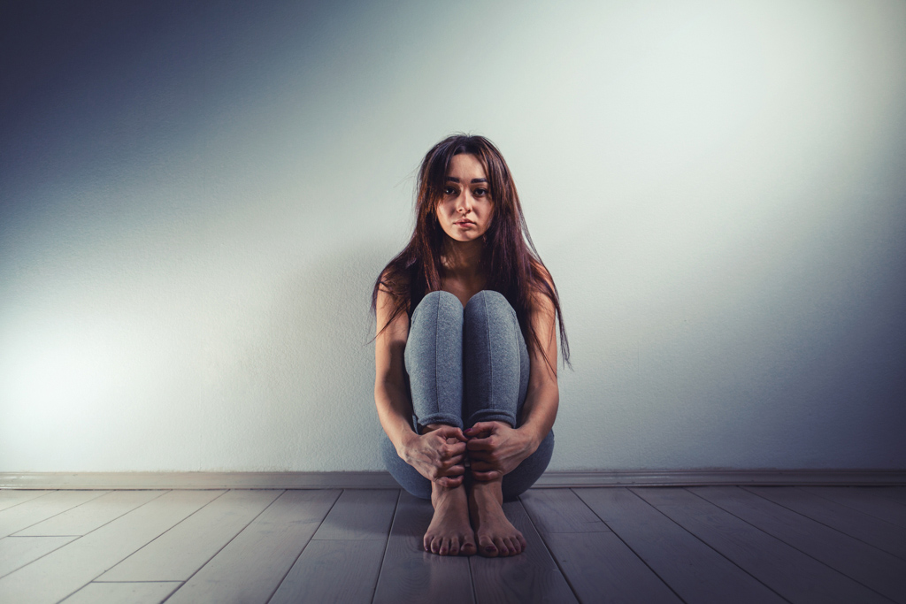 A young girl sitting on the floor alone looking alone with her hands around her ankles.