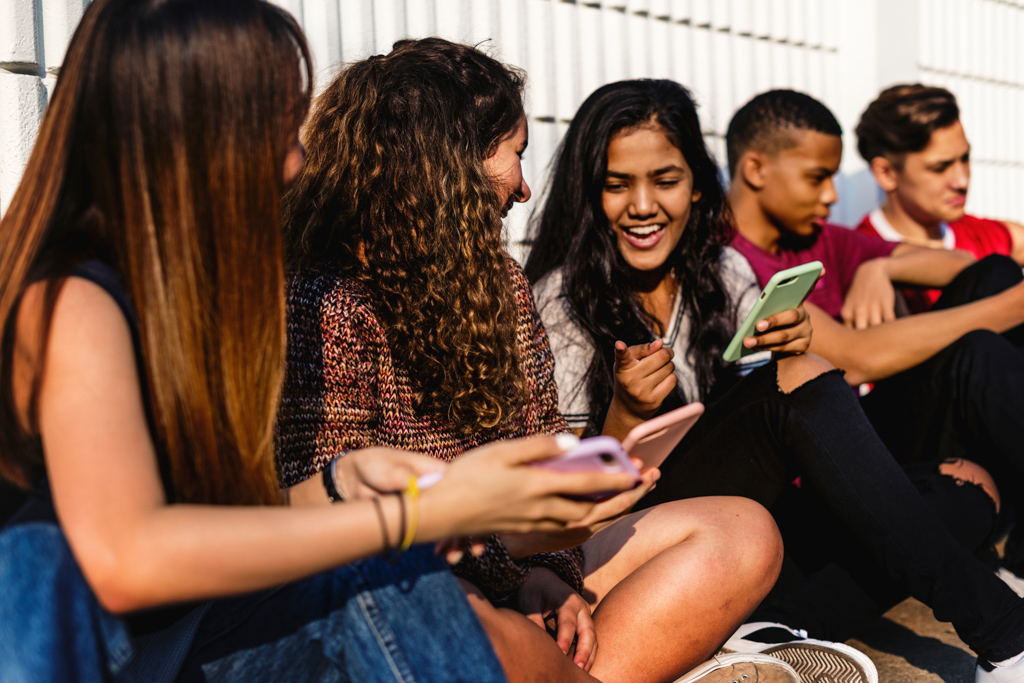 Five young adults sitting against a fence, one of the female young adults is laughing while holding her phone.