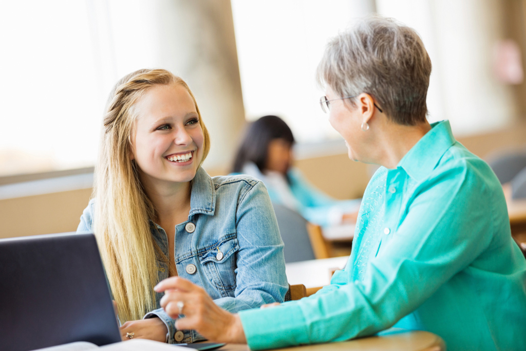 A woman and teen girl sitting at a table in front of a laptop, having a discussion and smiling.